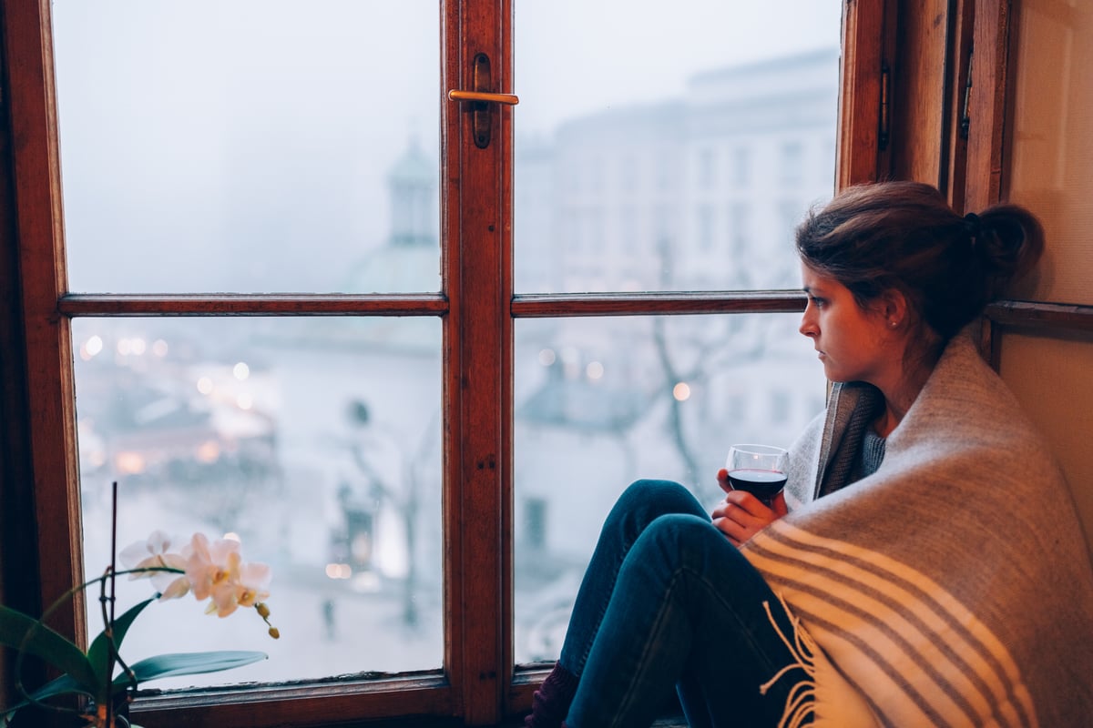 A worried woman wrapped in a blanket sits by a window.