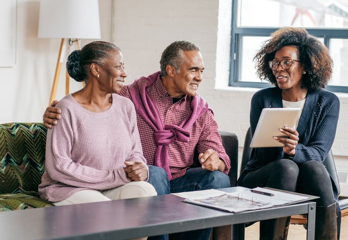 A senior couple sit on the couch talking to a real estate agent