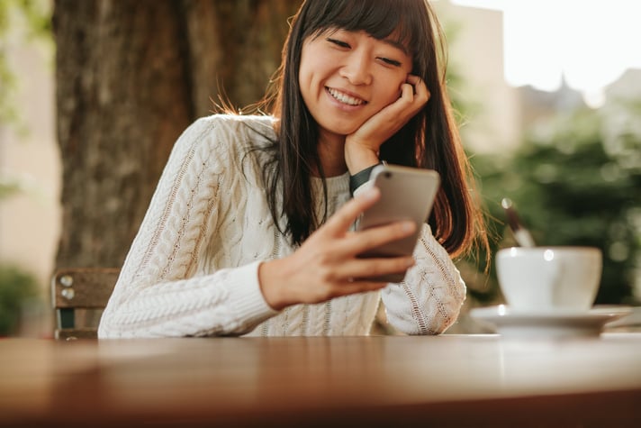 A smiling person sitting at an outdoor cafe table and looking at their cellphone.