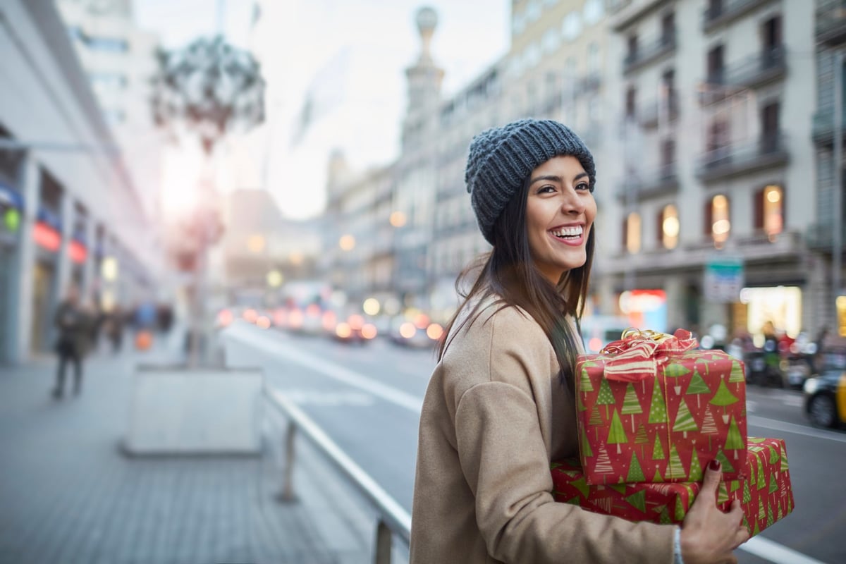 A young, smiling woman stands outside in the city with an armful of holiday gifts.