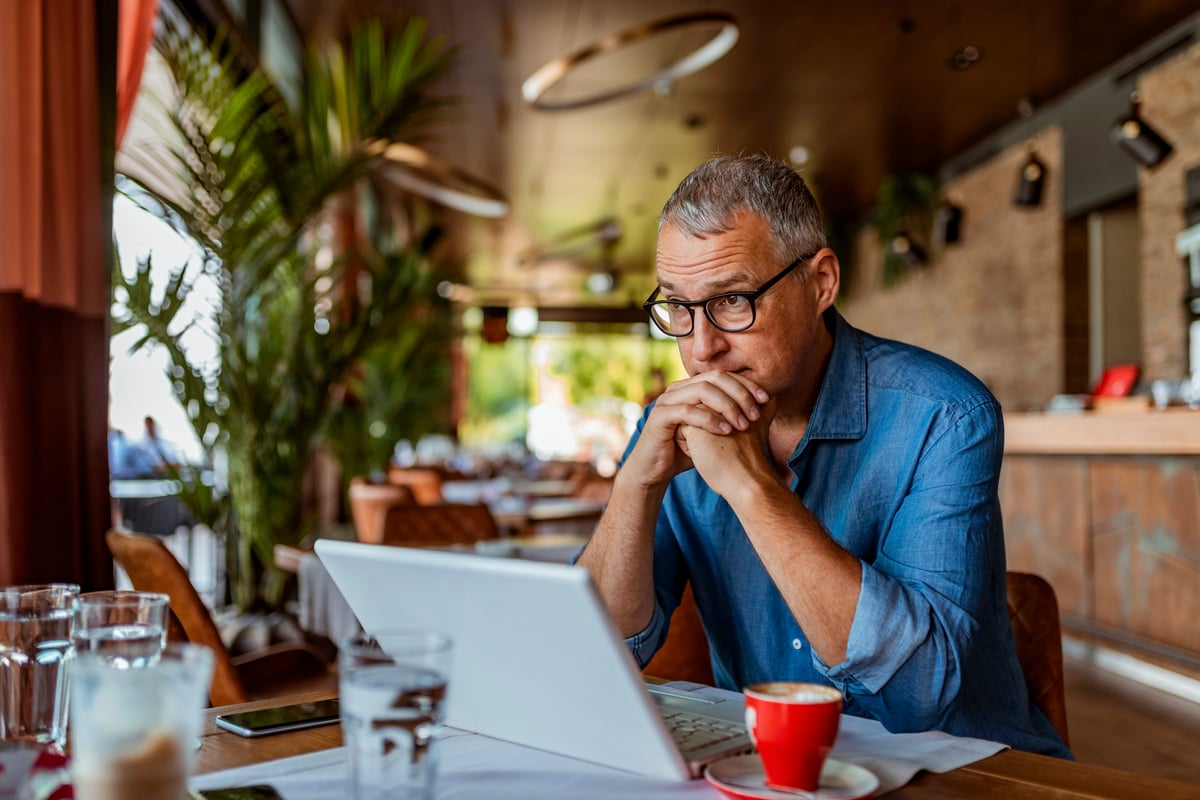 A man sitting in front of his laptop at a cafe table and looking stressed.