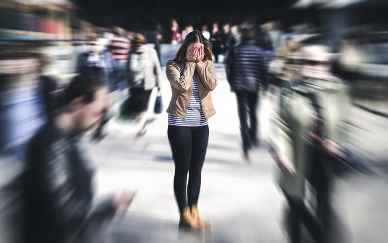 A stressed young woman in a busy crowd covering her face with her hands.