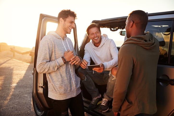 Three smiling men standing around their car and planning their route on a phone.