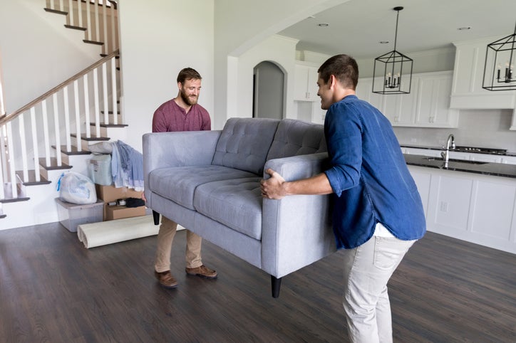 Two men smiling while carrying a couch into the living room of their new home.