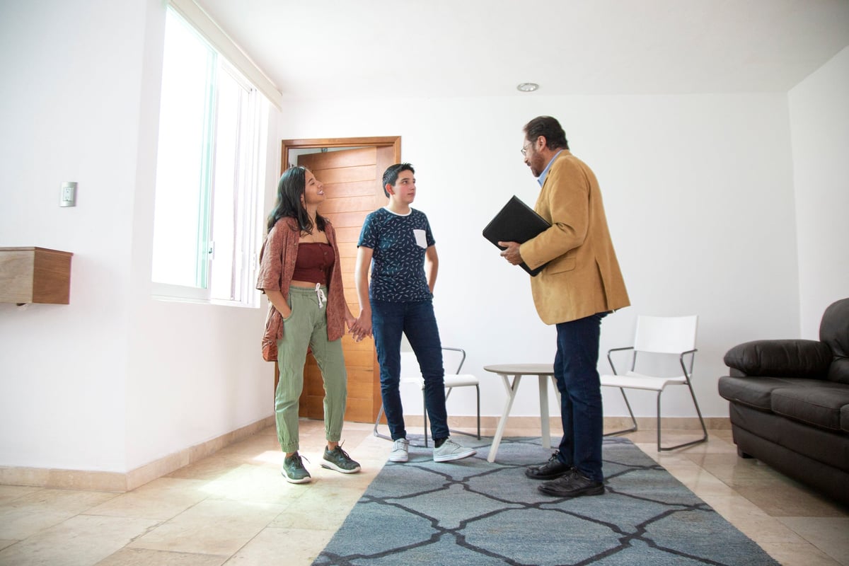 Two people holding hands while standing in the living room of a new house and talking with a realtor.