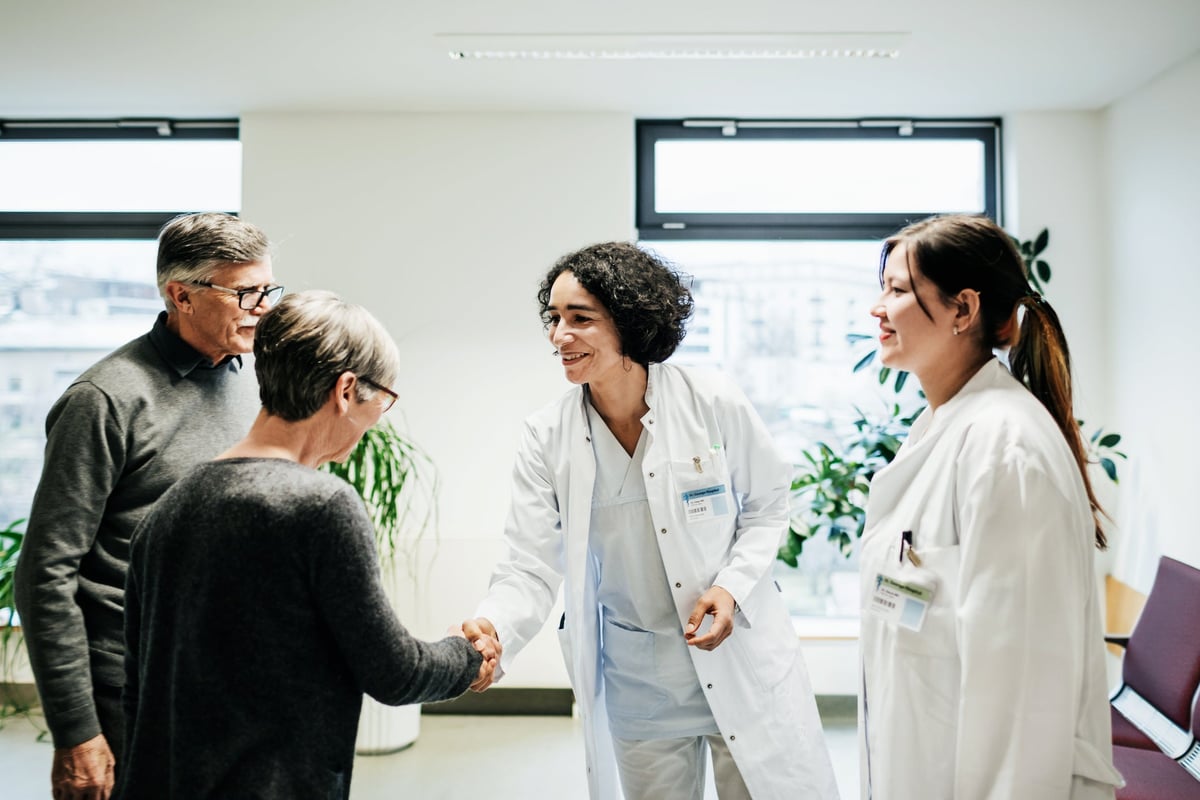 Two older people shaking hands with two smiling doctors in a sunny hospital room.