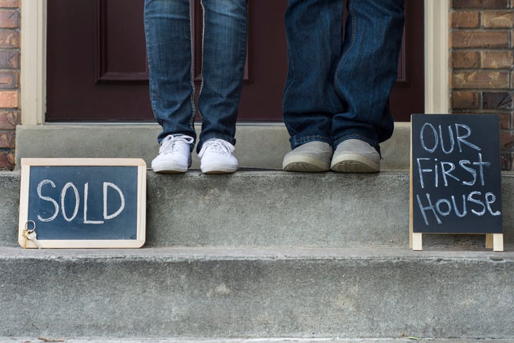 Two sets of feet standing on the front steps of a home next to signs saying Sold and Our First House.