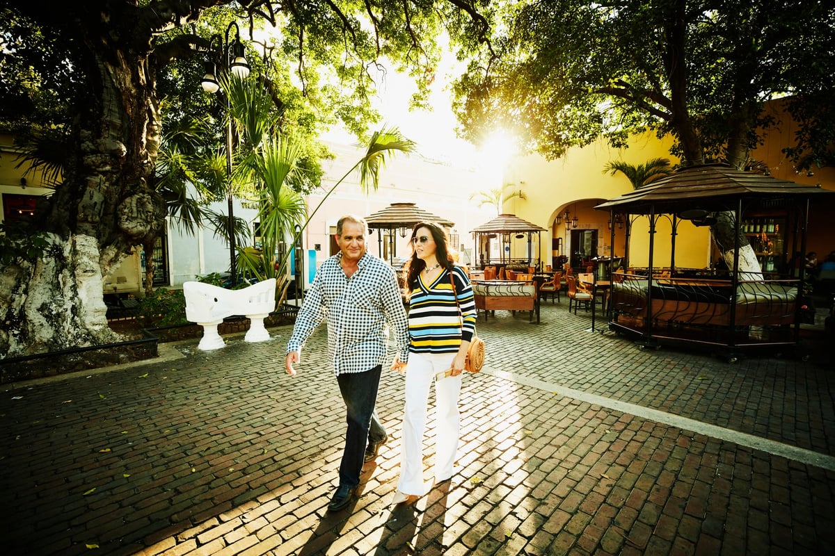 Two smiling people holding hands and walking through a sunny courtyard lined with trees.