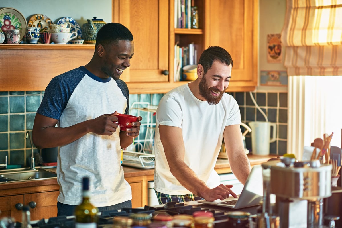 Two roommates laughing while watching something on a computer on the kitchen counter.