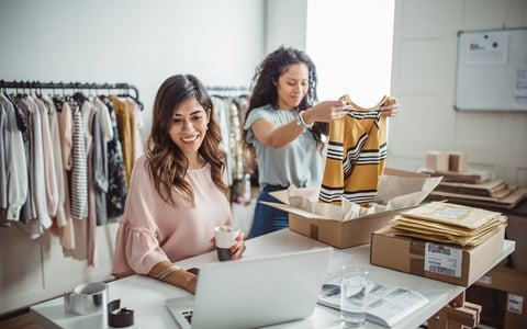 One woman packing clothes into shipping boxes while the other checks her laptop at a table in a clothing store storeroom.