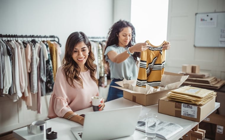 One woman packing clothes into shipping boxes while the other checks her laptop at a table in a clothing store storeroom.