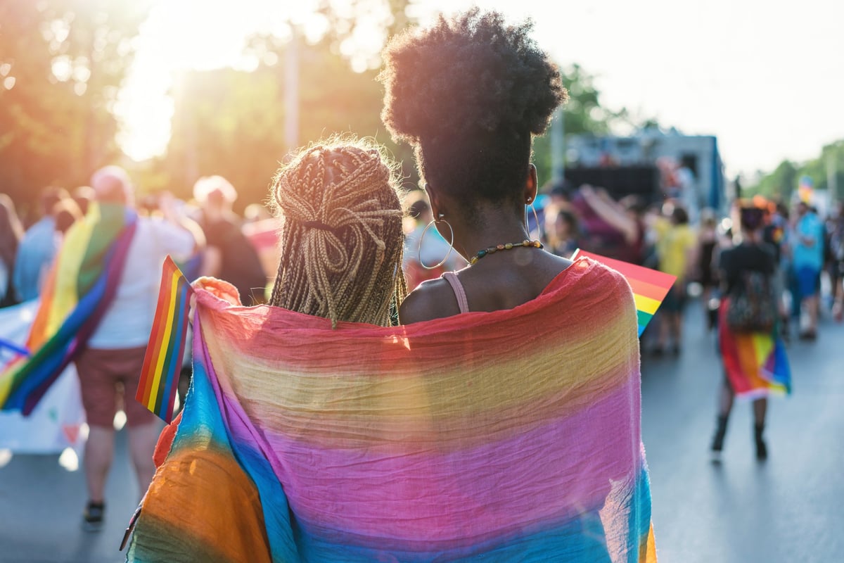 Two women wearing a rainbow flag around their shoulders at a Pride parade.