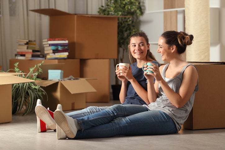 Two women sitting on the floor next to moving boxes while drinking coffee.