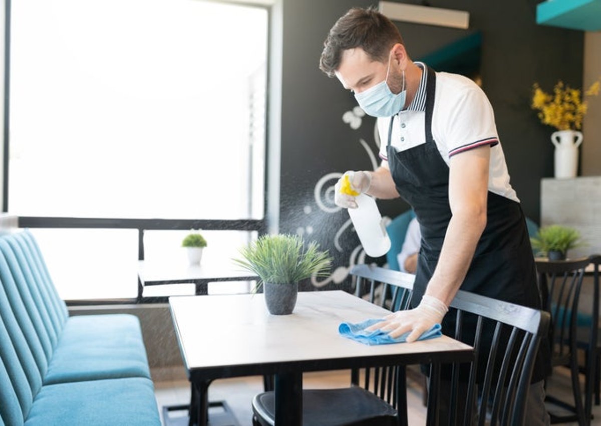 A waiter wearing a medical mask and cleaning a table in a restaurant with a spray bottle and cloth.