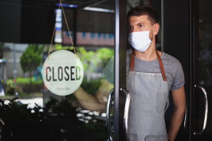 A waiter wearing a medical mask while standing in the doorway of an empty restaurant with a Closed sign hanging on the door.