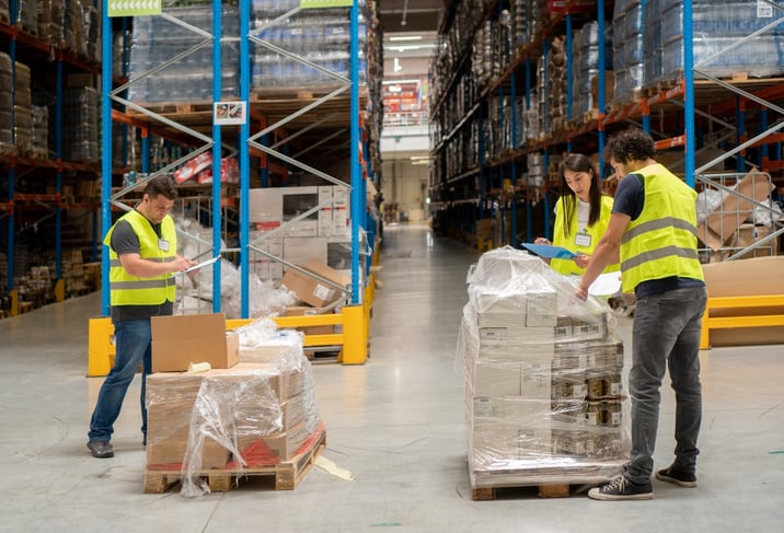 A woman and two men wearing safety vests unpack a pallet of boxes in a warehouse.