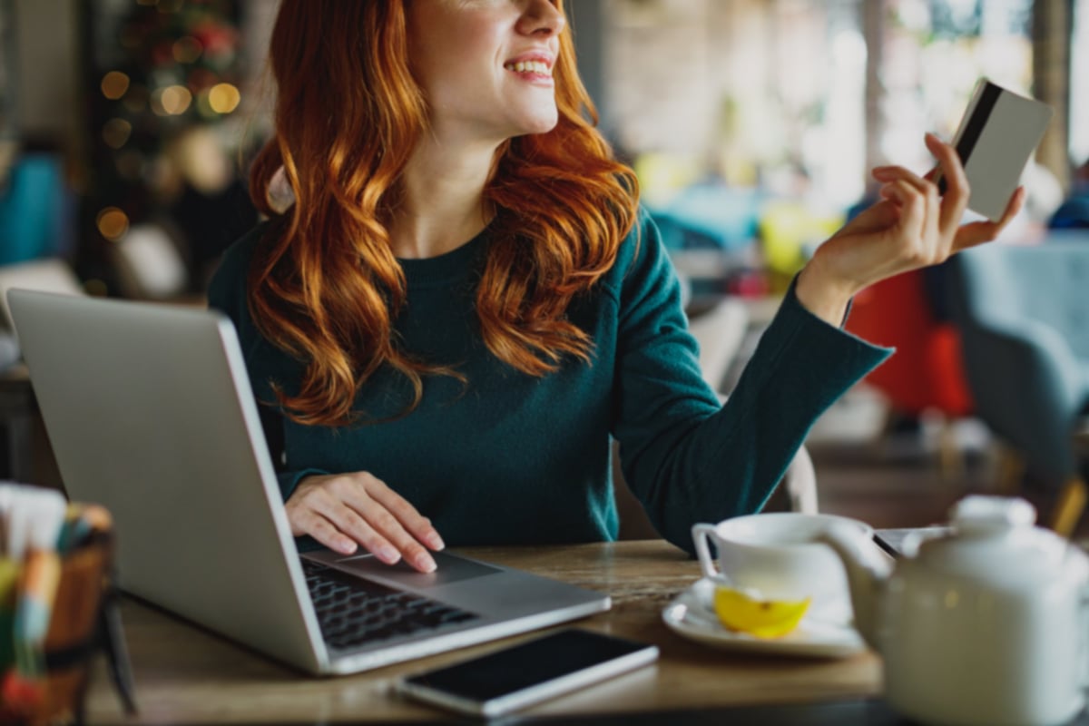 A woman sitting at a cafe table with tea and a laptop and holding out her credit card.