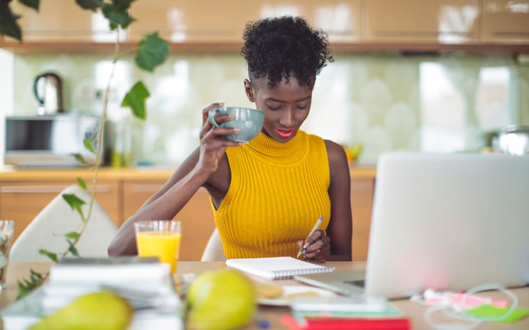 Woman drinking coffee at kitchen table while writing in a notebook with open laptop in front of her