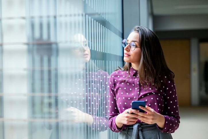 A young professional woman looking thoughtfully out an office window while holding a phone.