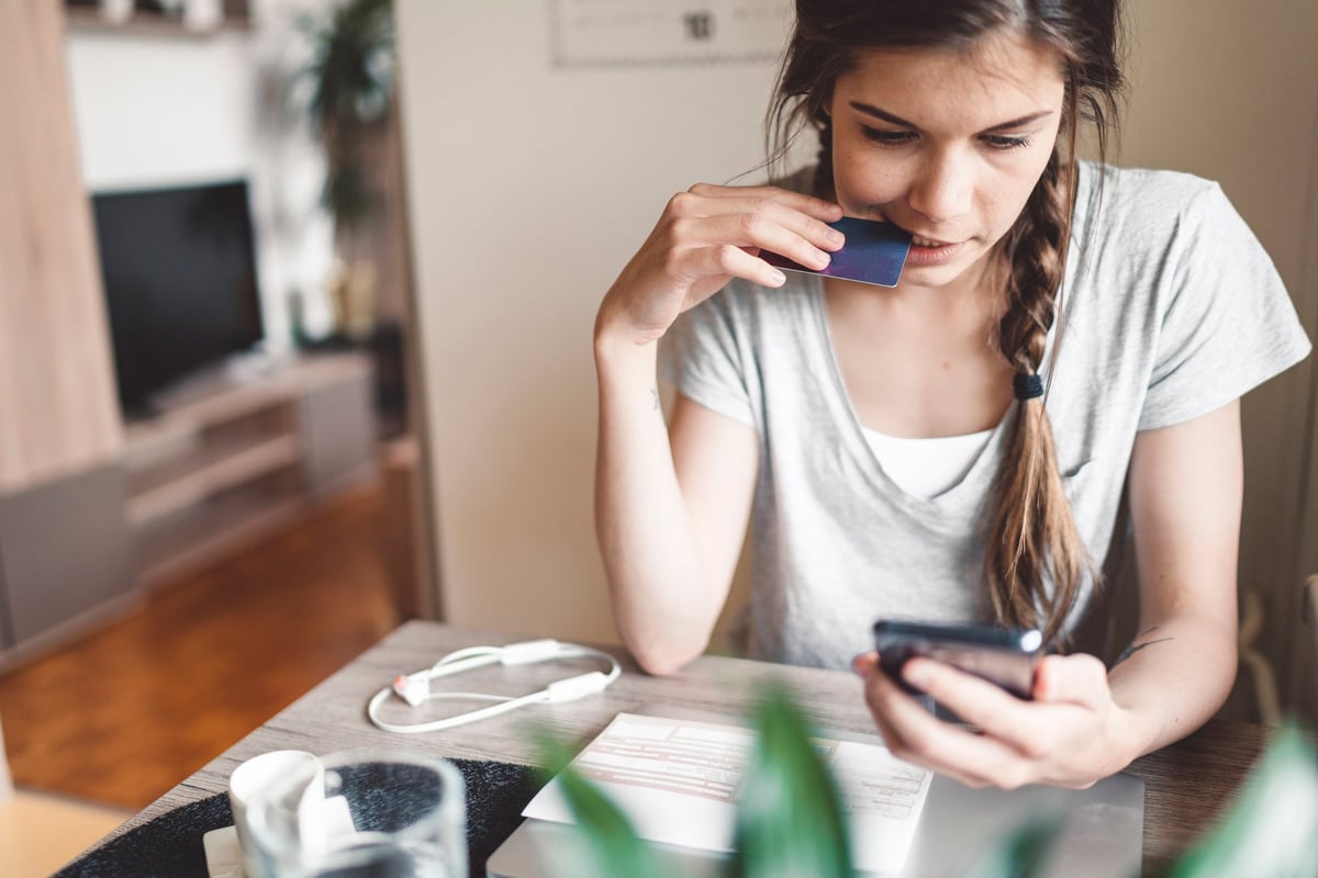 A young woman nervously holding a credit card up to her mouth while looking at her phone.