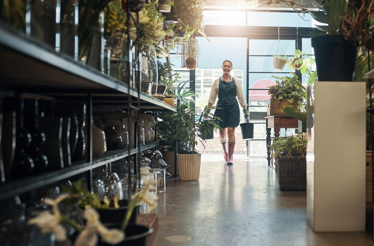 A woman in an apron and watering cans walks into her small shop lined with shelves of plants.