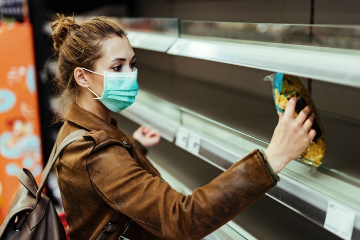 A woman wearing a medical mask and picking up the last bag of pasta from the grocery store shelf.