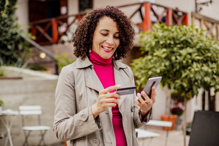 A smiling woman standing outside a sidewalk cafe looking at her phone while holding a credit card.