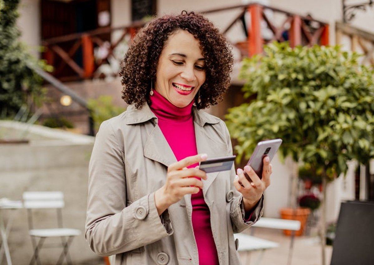 A smiling woman standing outside a sidewalk cafe looking at her phone while holding a credit card.