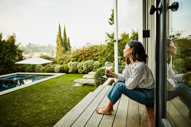 A smiling woman holding a glass of wine while seated on the steps of a patio deck looking at her backyard.