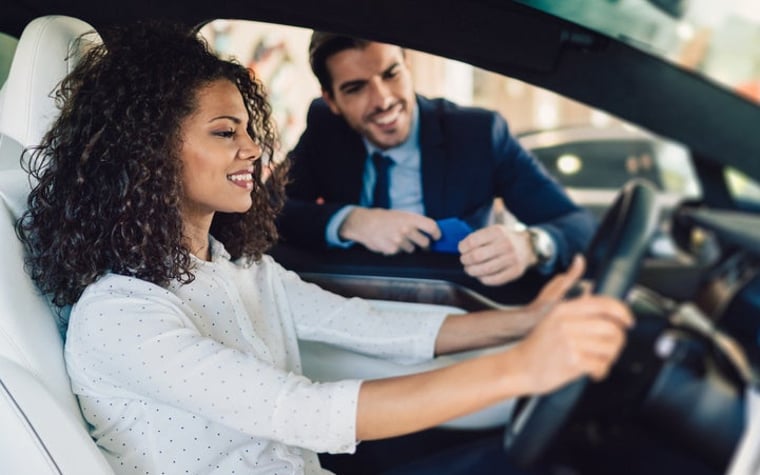A woman sitting in the driver's seat of a new car and talking to a salesman through the window.