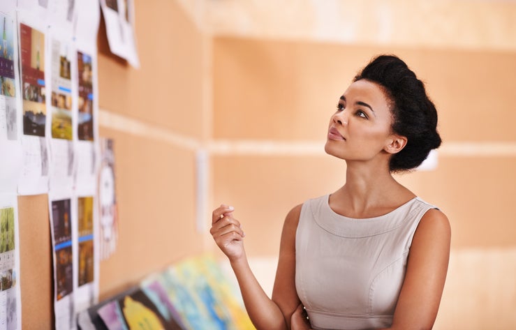 A young businesswoman looking thoughtfully at a wall of pictures in an office.