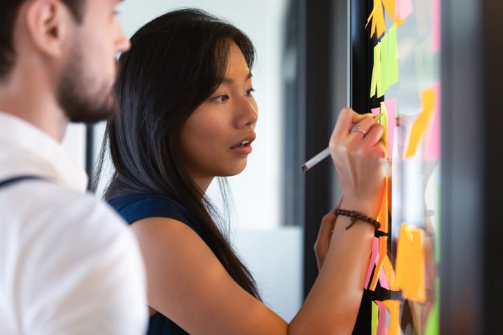 A woman in an office writing on sticky notes on a window while a man stands beside her.