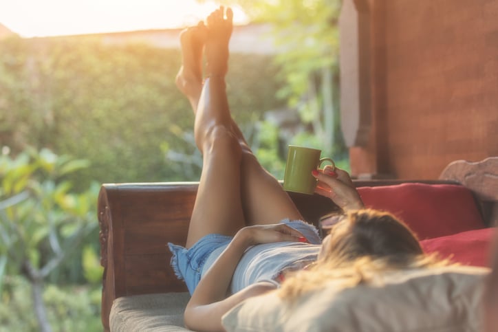 A woman lying down on a patio chair, looking at her sunny backyard, and holding a coffee mug.