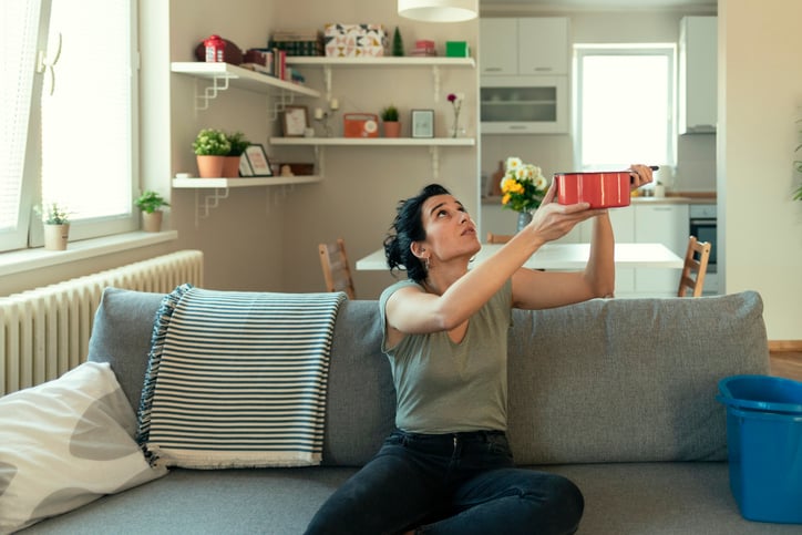 A woman sitting on her couch and holding up a pot to catch a leak falling from the ceiling.