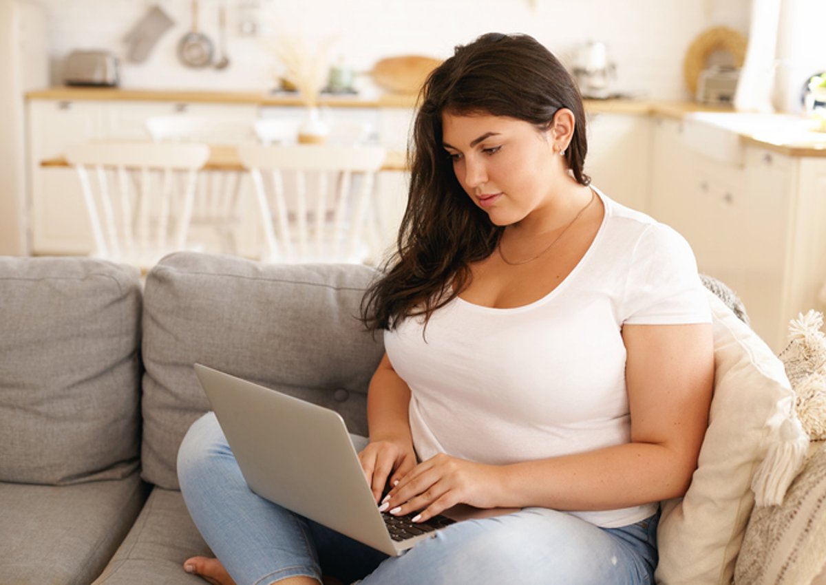 A woman sitting on the couch in her sunny living room and typing on her laptop.