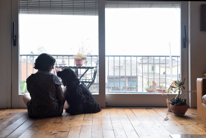 A worried woman sitting on the floor with her dog in front of her living room balcony window.