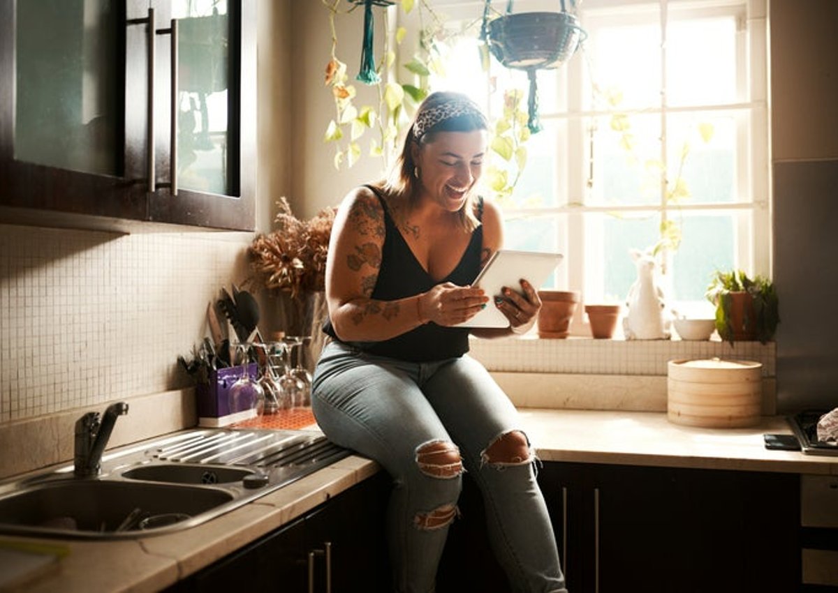 A woman sitting on the kitchen counter in her apartment and smiling at a tablet she's holding.