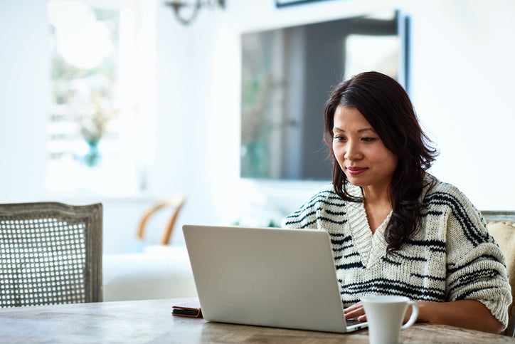 A woman sitting at her dining room table typing on her laptop.