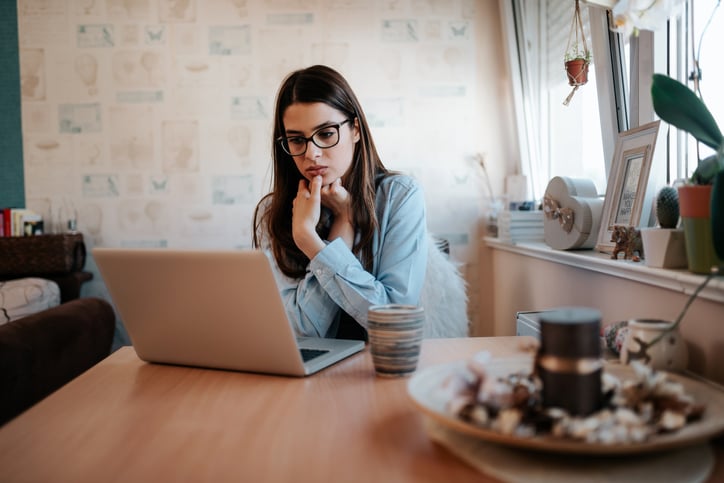 A worried-looking woman looking something up on her laptop while sitting at her kitchen table.