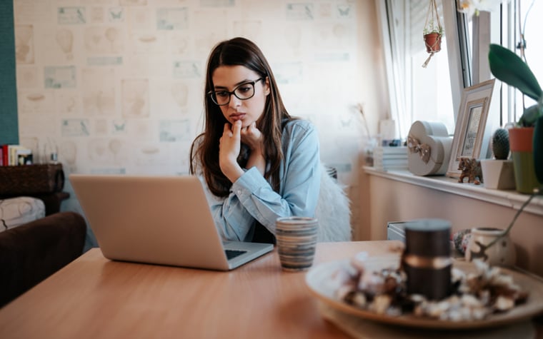 A worried-looking woman looking something up on her laptop while sitting at her kitchen table.