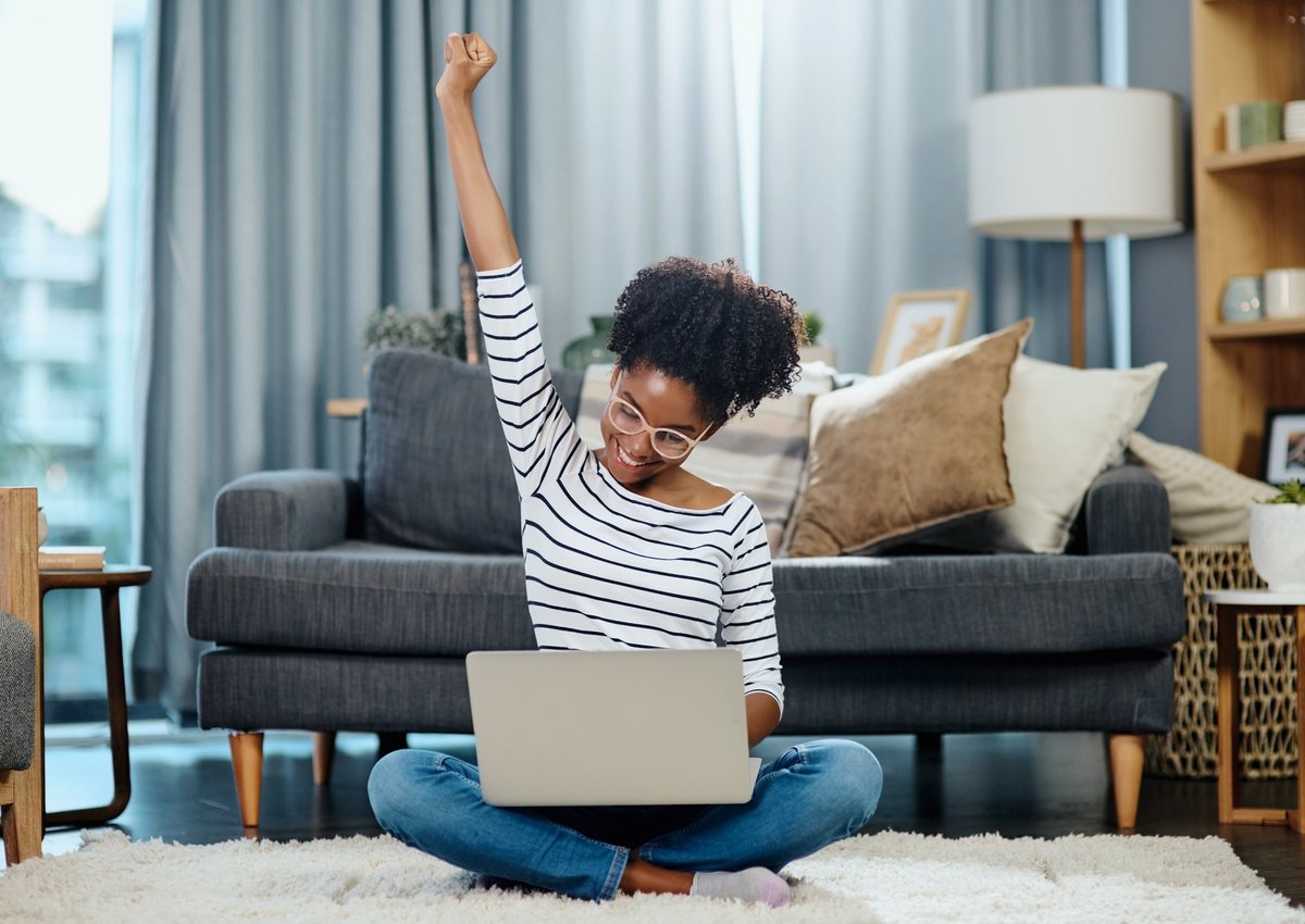 A woman sitting in front of her couch with her laptop and celebrating with her hand in the air.