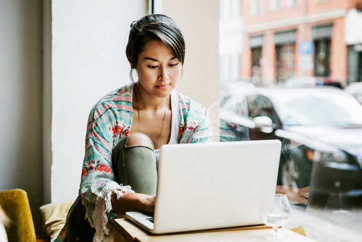 A woman sitting at a table by the window in a cafe and typing on a laptop.