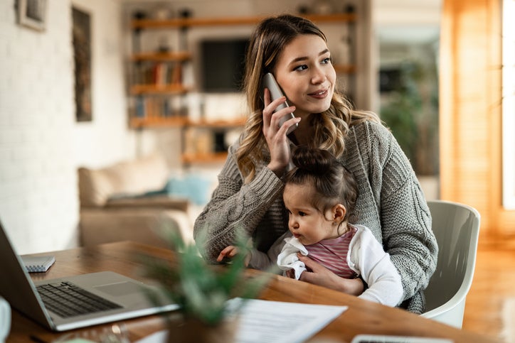 A woman sitting at a desk at home with her baby in her lap while making a phone call.