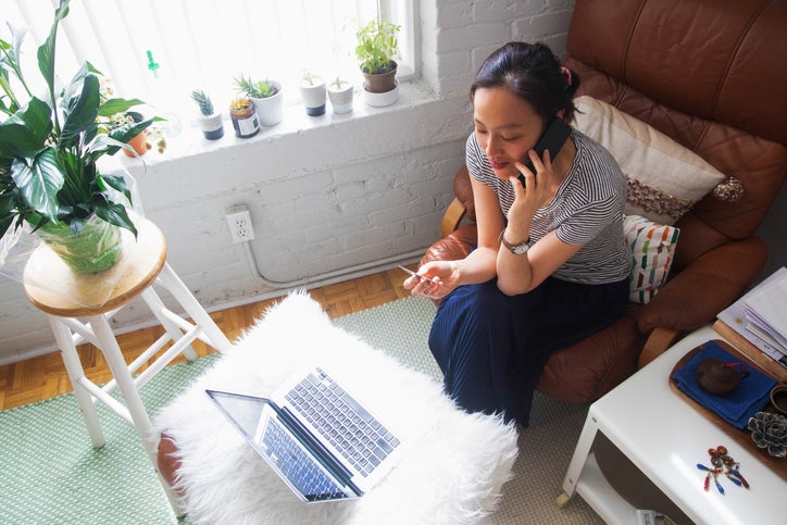 A woman sitting on her couch at home making a phone call while holding a credit card with a laptop open in front of her.