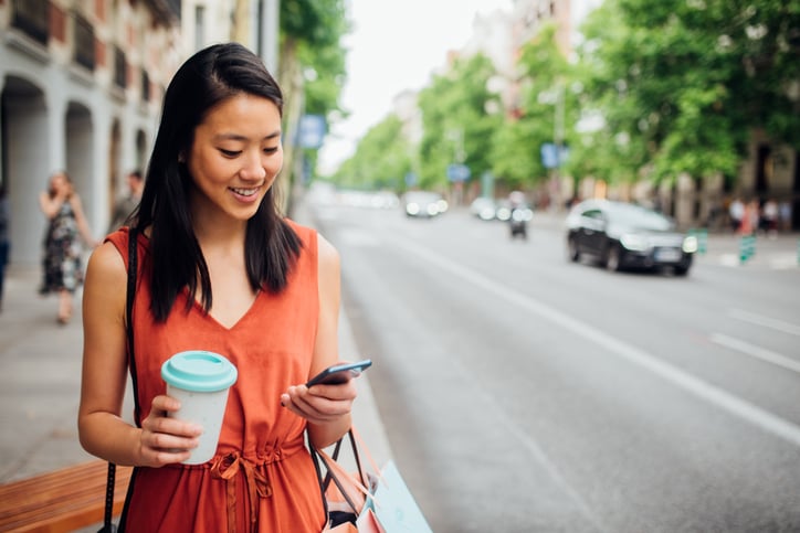 A girl walking down the street with a coffee in one hand and looking at her phone in the other hand.