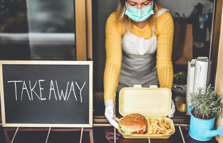 A woman wearing a medical mask handing a burger and fries through a window next to a sign that says Takeaway.