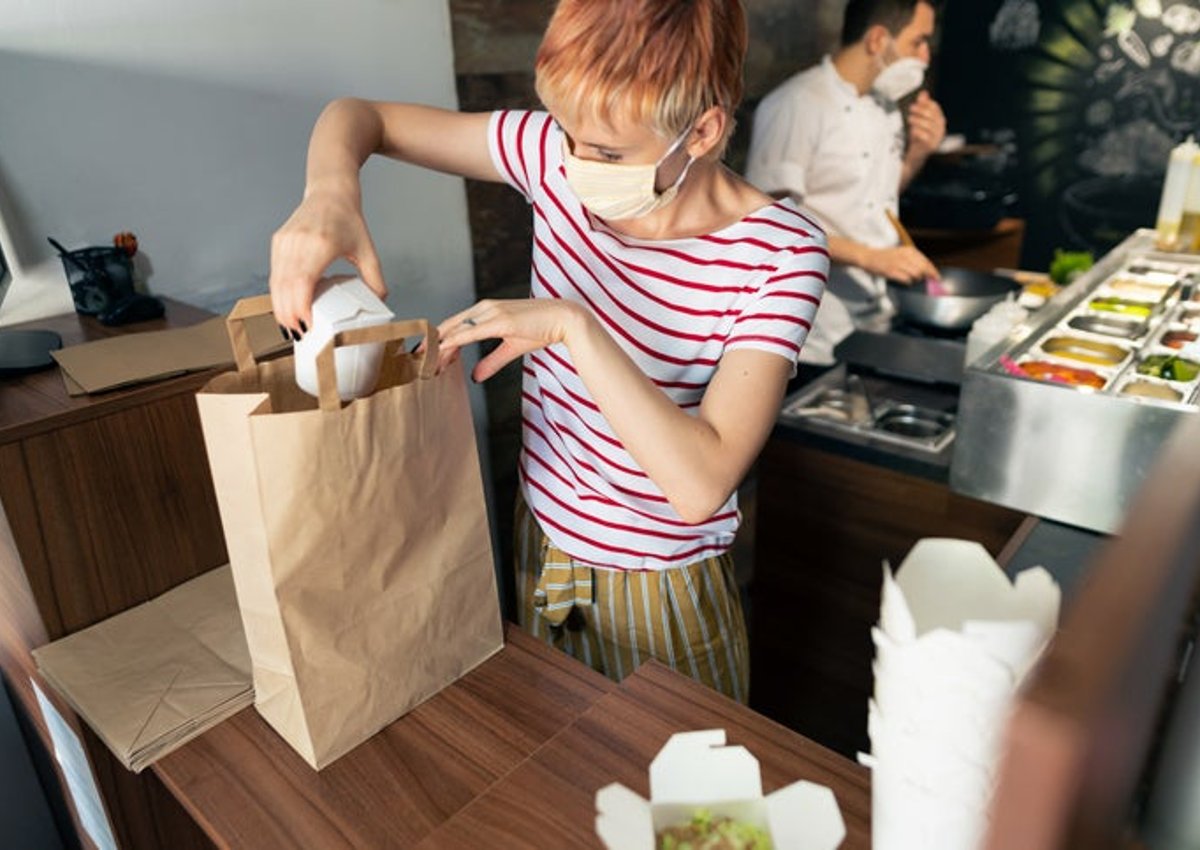 A person wearing a medical mask while packing takeout food containers into a bag with a chef working behind them.
