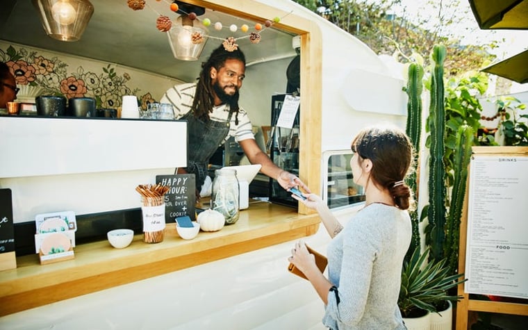 A woman hands her credit card through the window of a food truck to the owner standing inside wearing an apron.