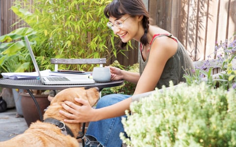 A person petting a dog while sitting outside at a patio table with an open laptop and a cup of coffee.