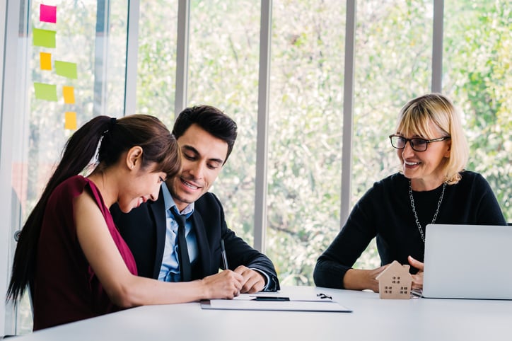 A smiling woman is signing documents while sitting at a table with her husband and his real estate agent.